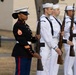 A formal military ceremony featuring a U.S. Marine Corps female staff sergeant in dress blues standing at attention alongside U.S. Navy sailors in dress whites. All personnel are holding rifles in a ceremonial position, suggesting a joint-service honor guard or parade formation. The setting appears to be an outdoor venue, likely a military installation.