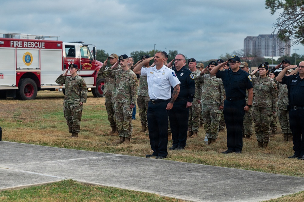 A formal outdoor ceremony showing military personnel in camouflage uniforms and civilian first responders rendering salutes together. A Fire Rescue truck bearing a United States Air Force emblem is visible in the background, suggesting this is a military installation ceremony. The gathering appears to be a memorial or commemorative event involving both military and civilian emergency services personnel.