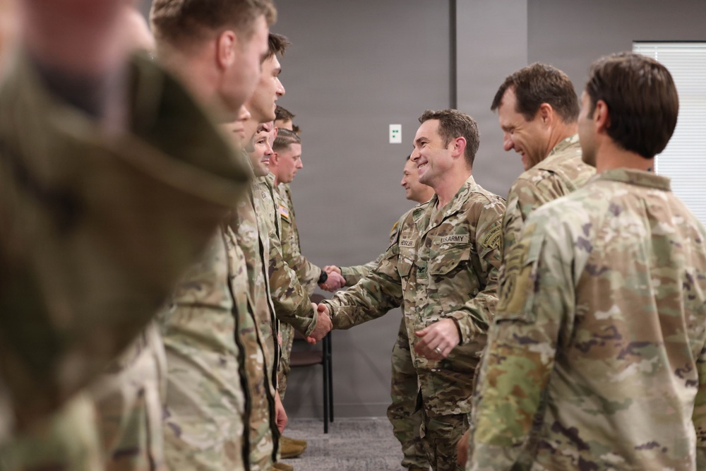 A group of U.S. Army soldiers in OCP (Operational Camouflage Pattern) uniforms are engaged in a formal greeting or recognition ceremony indoors. A central figure wearing a 'US ARMY' name tape is shaking hands with soldiers lined up in a row. The atmosphere appears positive, with visible smiling among the participants.