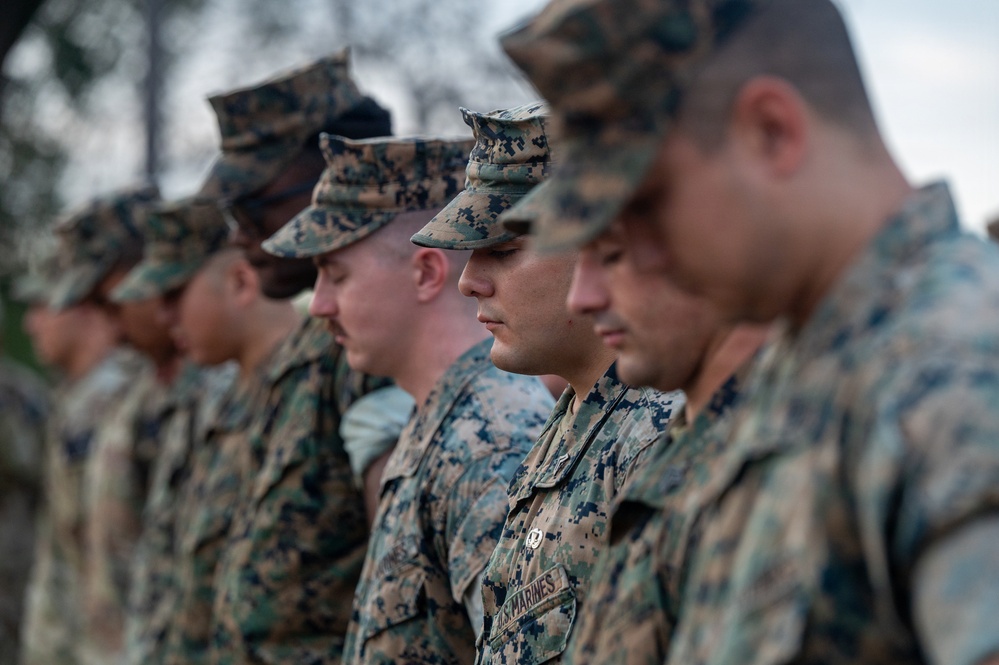 A group of U.S. Marines standing in formation, wearing MARPAT (Marine Pattern) digital camouflage uniforms and covers (caps). The Marines appear to be at attention or in a moment of reflection, with heads slightly bowed. The image is taken from a side angle with shallow depth of field, keeping the middle Marines in sharpest focus.