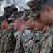 A group of U.S. Marines standing in formation, wearing MARPAT (Marine Pattern) digital camouflage uniforms and covers (caps). The Marines appear to be at attention or in a moment of reflection, with heads slightly bowed. The image is taken from a side angle with shallow depth of field, keeping the middle Marines in sharpest focus.