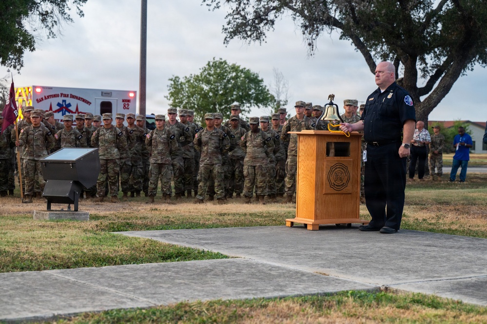 A uniformed fire department official stands at a wooden podium bearing a ceremonial bell, addressing a large formation of military personnel in camouflage uniforms. The setting appears to be an outdoor memorial or remembrance ceremony, likely a 9/11 commemoration given the ceremonial bell and formal military formation. A San Antonio Fire Department ambulance is visible in the background.