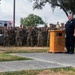 A uniformed fire department official stands at a wooden podium bearing a ceremonial bell, addressing a large formation of military personnel in camouflage uniforms. The setting appears to be an outdoor memorial or remembrance ceremony, likely a 9/11 commemoration given the ceremonial bell and formal military formation. A San Antonio Fire Department ambulance is visible in the background.