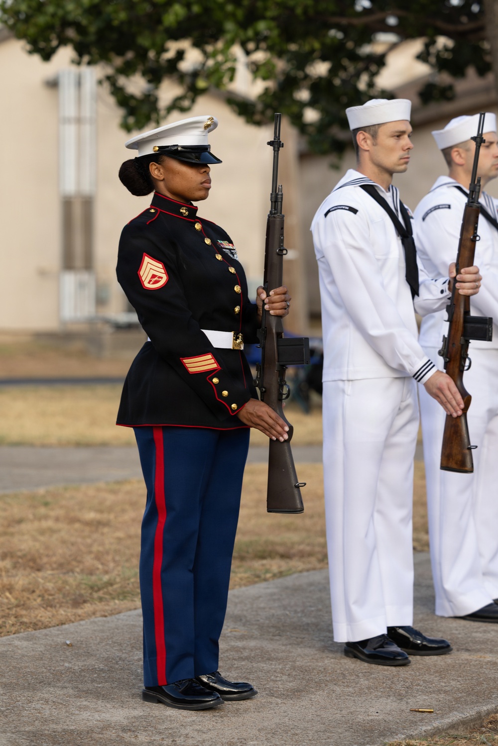 A U.S. Marine in dress blues stands at attention holding a rifle during what appears to be a formal military ceremony. Behind her, two U.S. Navy sailors in dress whites also stand at attention with rifles. The setting appears to be an outdoor ceremonial event on a military installation.