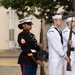 A U.S. Marine in dress blues stands at attention holding a rifle during what appears to be a formal military ceremony. Behind her, two U.S. Navy sailors in dress whites also stand at attention with rifles. The setting appears to be an outdoor ceremonial event on a military installation.