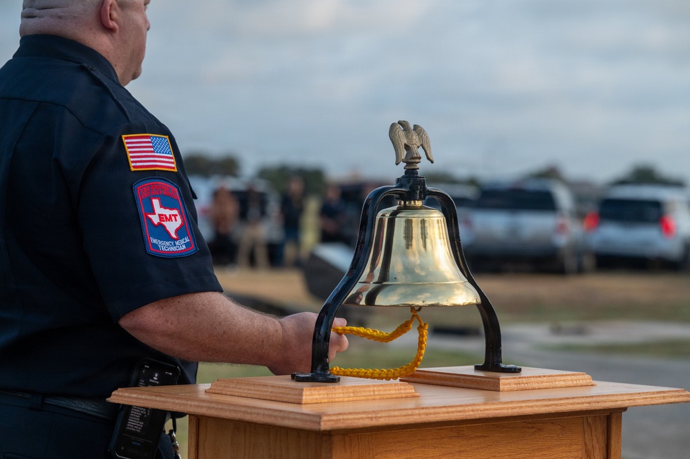 A Texas EMT (Emergency Medical Technician) stands beside a ceremonial brass bell mounted on a wooden podium during what appears to be a memorial or remembrance ceremony. The bell, topped with an eagle figurine and fitted with a yellow braided pull cord, is a traditional symbol used in first responder memorial services. A group of people can be seen gathered in the background, suggesting a formal outdoor ceremony.