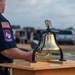 A Texas EMT (Emergency Medical Technician) stands beside a ceremonial brass bell mounted on a wooden podium during what appears to be a memorial or remembrance ceremony. The bell, topped with an eagle figurine and fitted with a yellow braided pull cord, is a traditional symbol used in first responder memorial services. A group of people can be seen gathered in the background, suggesting a formal outdoor ceremony.