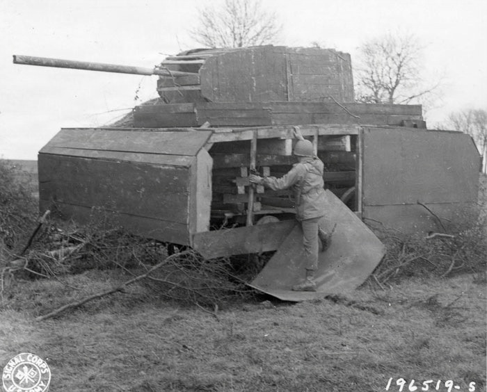 A U.S. Army Signal Corps photograph (number 196519-S) showing a wooden dummy tank decoy used for military deception purposes. A soldier in uniform and helmet is examining or adjusting the interior mechanical components visible through an open panel on the side of the fake tank. The structure is constructed from flat wooden panels shaped to resemble a tank hull and turret, with a pipe serving as a mock gun barrel.