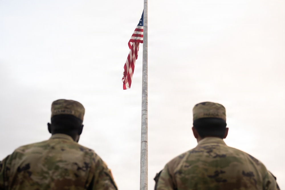 Two U.S. military personnel in Army Combat Uniforms (ACU) stand at attention facing an American flag on a flagpole. The flag appears to be positioned at or near half-staff, suggesting a memorial or mourning observance. The overcast sky and rear-facing composition give the image a solemn, ceremonial tone.