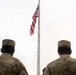 Two U.S. military personnel in Army Combat Uniforms (ACU) stand at attention facing an American flag on a flagpole. The flag appears to be positioned at or near half-staff, suggesting a memorial or mourning observance. The overcast sky and rear-facing composition give the image a solemn, ceremonial tone.