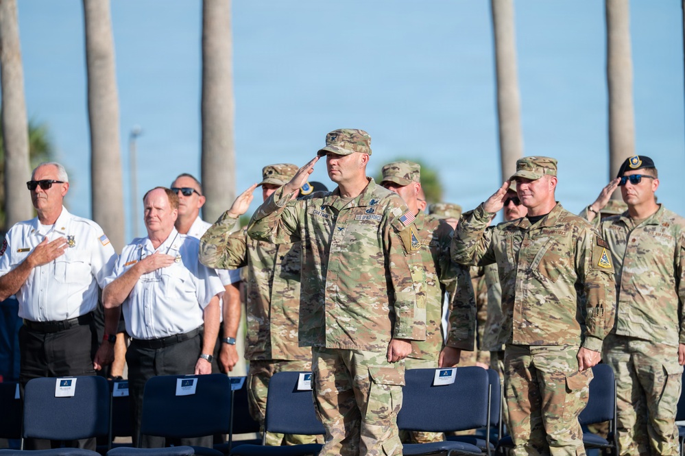 A formal military and civilian ceremony is taking place outdoors, likely a memorial or patriotic event. Military personnel in OCP (Operational Camouflage Pattern) uniforms are rendering salutes while civilians in white uniform shirts place their hands over their hearts. The setting features palm trees in the background and rows of blue chairs in the foreground.