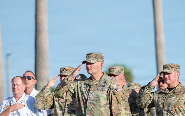 A formal military and civilian ceremony is taking place outdoors, likely a memorial or patriotic event. Military personnel in OCP (Operational Camouflage Pattern) uniforms are rendering salutes while civilians in white uniform shirts place their hands over their hearts. The setting features palm trees in the background and rows of blue chairs in the foreground.