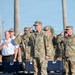 A formal military and civilian ceremony is taking place outdoors, likely a memorial or patriotic event. Military personnel in OCP (Operational Camouflage Pattern) uniforms are rendering salutes while civilians in white uniform shirts place their hands over their hearts. The setting features palm trees in the background and rows of blue chairs in the foreground.