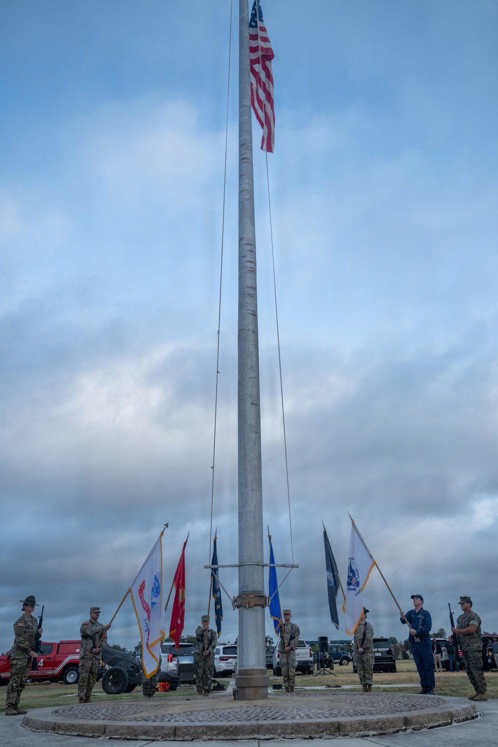 A military color guard ceremony is taking place around a large flagpole with the American flag flying at the top. Multiple service members in camouflage uniforms are holding various military branch flags surrounding the base of the flagpole. The ceremony appears to be an outdoor formal military event with vehicles and equipment visible in the background.