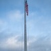 A military color guard ceremony is taking place around a large flagpole with the American flag flying at the top. Multiple service members in camouflage uniforms are holding various military branch flags surrounding the base of the flagpole. The ceremony appears to be an outdoor formal military event with vehicles and equipment visible in the background.