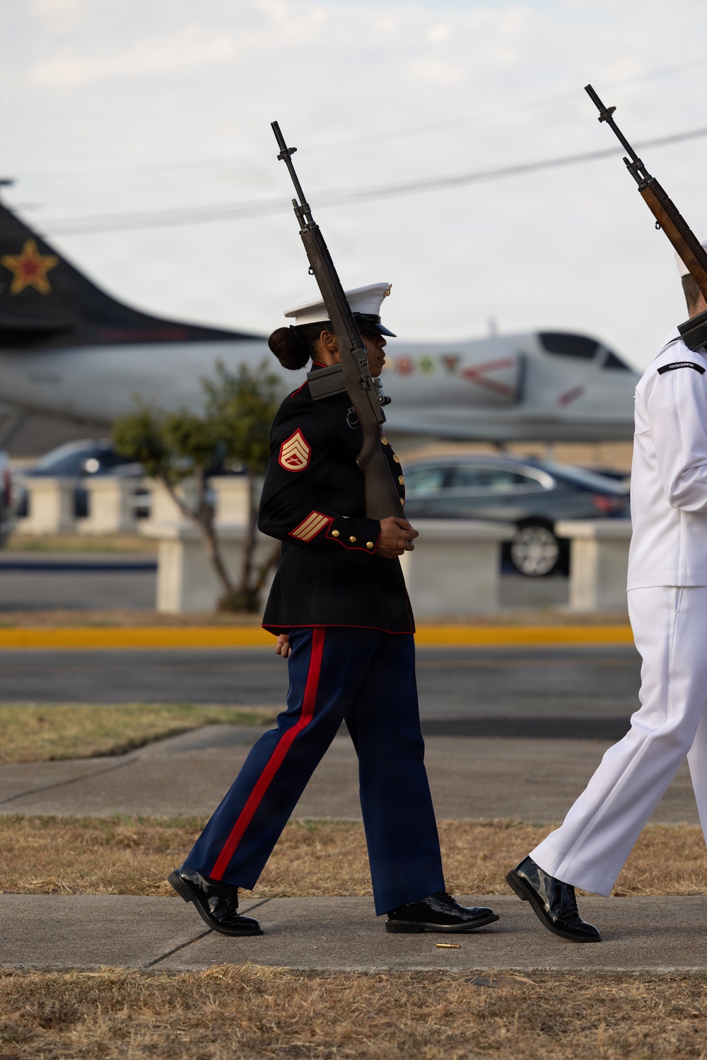 A U.S. Marine Corps Staff Sergeant in dress blues uniform marches in a ceremonial formation carrying an M14 rifle at port arms. A U.S. Navy sailor in dress whites is visible to the right, also carrying a rifle. The setting appears to be a military base or airfield, with aircraft visible in the background.