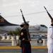 A U.S. Marine Corps Staff Sergeant in dress blues uniform marches in a ceremonial formation carrying an M14 rifle at port arms. A U.S. Navy sailor in dress whites is visible to the right, also carrying a rifle. The setting appears to be a military base or airfield, with aircraft visible in the background.