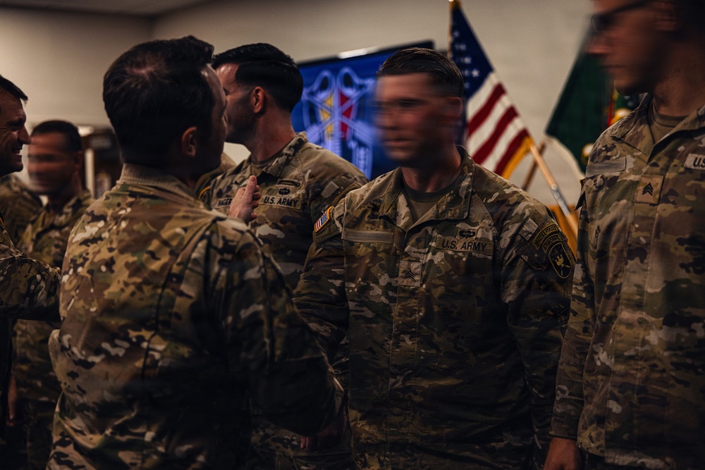 A group of U.S. Army soldiers in Operational Camouflage Pattern (OCP) uniforms are gathered in what appears to be an indoor military ceremony or formation. Several soldiers are interacting closely, possibly during a pinning ceremony, award presentation, or unit formation event. An American flag and what appears to be a unit guidon or organizational flag are visible in the background.
