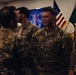 A group of U.S. Army soldiers in Operational Camouflage Pattern (OCP) uniforms are gathered in what appears to be an indoor military ceremony or formation. Several soldiers are interacting closely, possibly during a pinning ceremony, award presentation, or unit formation event. An American flag and what appears to be a unit guidon or organizational flag are visible in the background.