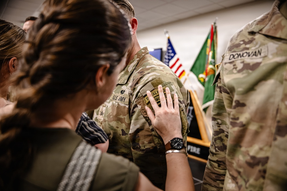 A U.S. Army military ceremony is taking place indoors, likely a promotion or pinning event. A woman is placing her hand on the shoulder/chest area of a soldier in OCP (Operational Camouflage Pattern) uniform, suggesting a rank pinning or badge placement. A second soldier identified by the name tape 'DONOVAN' is visible on the right side of the frame.