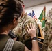 A U.S. Army military ceremony is taking place indoors, likely a promotion or pinning event. A woman is placing her hand on the shoulder/chest area of a soldier in OCP (Operational Camouflage Pattern) uniform, suggesting a rank pinning or badge placement. A second soldier identified by the name tape 'DONOVAN' is visible on the right side of the frame.