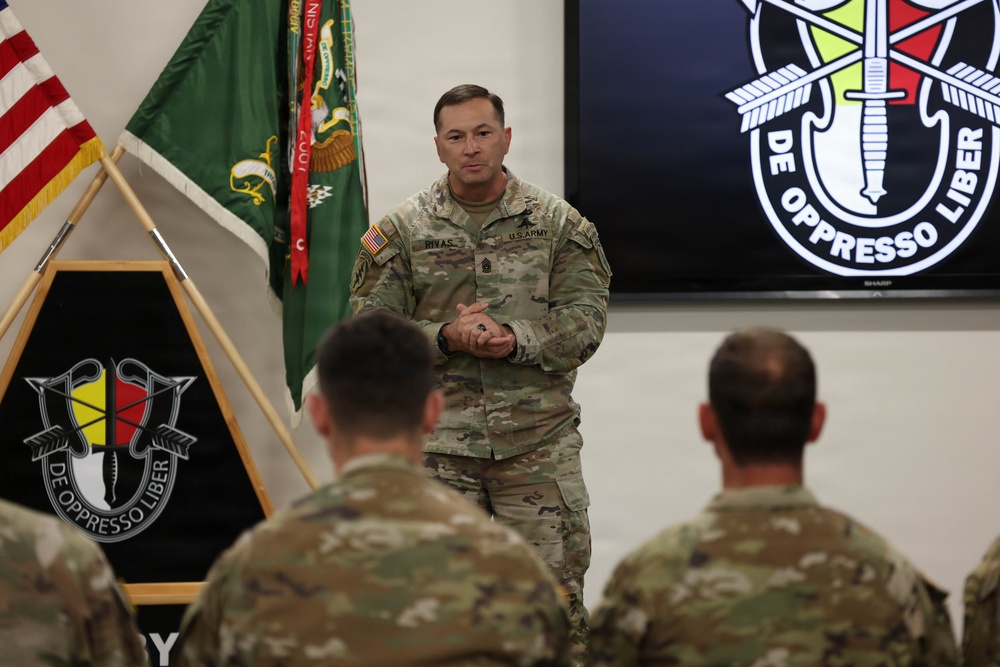 A U.S. Army senior non-commissioned officer named Rivas is addressing a group of soldiers in a formal military setting. The backdrop features the U.S. Army Special Forces insignia and motto 'De Oppresso Liber,' indicating this is a Special Forces unit gathering. The speaker appears to be conducting a briefing or address to assembled personnel.