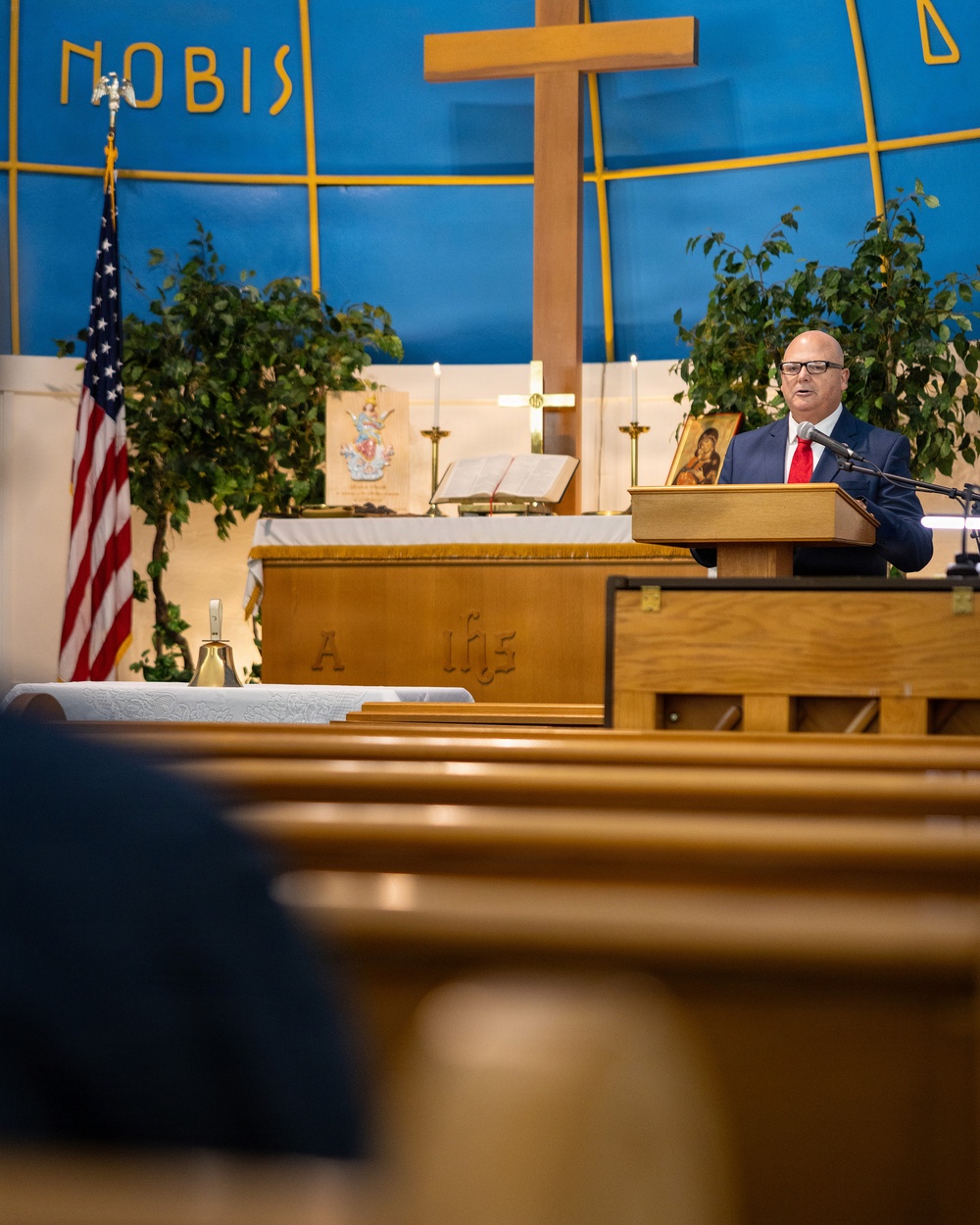 A man in a dark suit with a red tie is speaking at a wooden podium/lectern inside a Christian church. The sanctuary features a large wooden cross, blue and gold wall decorations with the partial text 'NOBIS', an American flag, green plants, candles, and religious iconography. The perspective is shot from a low angle through the church pews toward the altar area.