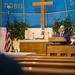 A man in a dark suit with a red tie is speaking at a wooden podium/lectern inside a Christian church. The sanctuary features a large wooden cross, blue and gold wall decorations with the partial text 'NOBIS', an American flag, green plants, candles, and religious iconography. The perspective is shot from a low angle through the church pews toward the altar area.