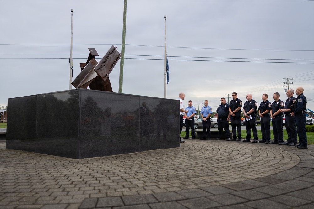 A group of uniformed first responders, primarily firefighters and emergency personnel, are gathered at an outdoor memorial featuring a dark granite base with twisted steel beam sculpture, consistent with 9/11 memorial designs. The flags in the background appear to be at half-staff, suggesting a commemorative ceremony. A civilian in a white shirt appears to be addressing or greeting the assembled personnel.