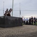 A group of uniformed first responders, primarily firefighters and emergency personnel, are gathered at an outdoor memorial featuring a dark granite base with twisted steel beam sculpture, consistent with 9/11 memorial designs. The flags in the background appear to be at half-staff, suggesting a commemorative ceremony. A civilian in a white shirt appears to be addressing or greeting the assembled personnel.