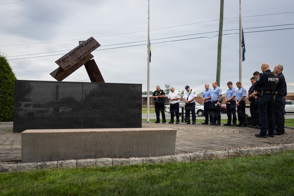 A group of uniformed first responders, including police officers and firefighters, stand in a semicircle with heads bowed in a moment of silence or prayer at an outdoor memorial. The memorial features twisted steel beam fragments mounted on a dark granite base, consistent with 9/11 memorial structures found across the United States. The ceremony takes place on a brick plaza with flagpoles visible in the background under an overcast sky.