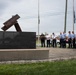 A group of uniformed first responders, including police officers and firefighters, stand in a semicircle with heads bowed in a moment of silence or prayer at an outdoor memorial. The memorial features twisted steel beam fragments mounted on a dark granite base, consistent with 9/11 memorial structures found across the United States. The ceremony takes place on a brick plaza with flagpoles visible in the background under an overcast sky.