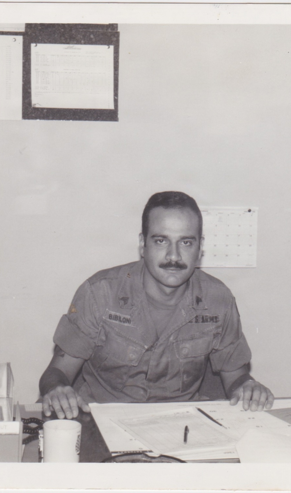 A black-and-white photograph of a U.S. Army soldier seated at a desk in what appears to be an office setting. The soldier, whose name tape reads 'BIBILONI' and whose right chest tape reads 'U.S. ARMY', is leaning forward over papers on the desk. Two calendars or charts are pinned to the wall behind him.