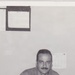 A black-and-white photograph of a U.S. Army soldier seated at a desk in what appears to be an office setting. The soldier, whose name tape reads 'BIBILONI' and whose right chest tape reads 'U.S. ARMY', is leaning forward over papers on the desk. Two calendars or charts are pinned to the wall behind him.
