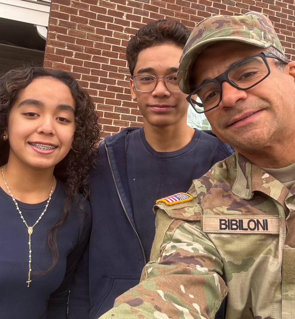 A selfie photograph showing three individuals posing together in front of a brick wall. One adult male is wearing a U.S. Army OCP (Operational Camouflage Pattern) uniform with a name tape reading 'BIBILONI' and an American flag patch, accompanied by two younger individuals who appear to be teenagers. The image has the appearance of a family photo, likely taken during a military-related visit or event.