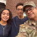 A selfie photograph showing three individuals posing together in front of a brick wall. One adult male is wearing a U.S. Army OCP (Operational Camouflage Pattern) uniform with a name tape reading 'BIBILONI' and an American flag patch, accompanied by two younger individuals who appear to be teenagers. The image has the appearance of a family photo, likely taken during a military-related visit or event.