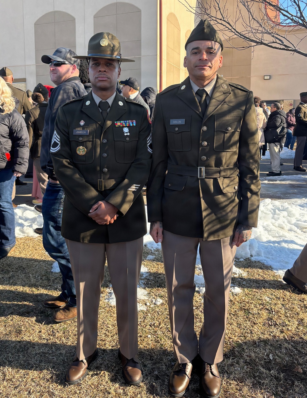 Two U.S. Army soldiers in Army Green Service Uniforms (Class A) are standing together outdoors, likely at a military ceremony or graduation event. The soldier on the left wears a drill sergeant hat and displays multiple service ribbons and a nameplate reading 'JOHNSON' with Staff Sergeant rank insignia. The soldier on the right wears a garrison cap with nameplate reading 'BIBILONI' and a belt with the older-style uniform configuration.