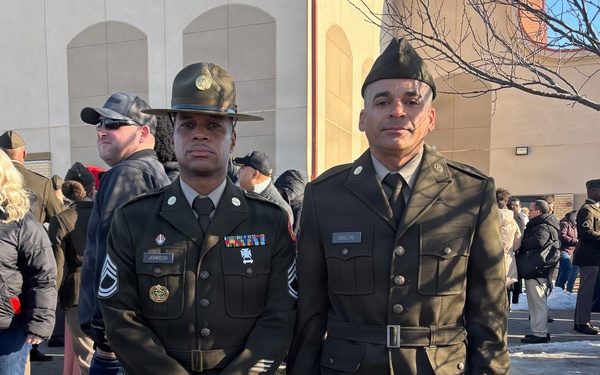 Two U.S. Army soldiers in Army Green Service Uniforms (Class A) are standing together outdoors, likely at a military ceremony or graduation event. The soldier on the left wears a drill sergeant hat and displays multiple service ribbons and a nameplate reading 'JOHNSON' with Staff Sergeant rank insignia. The soldier on the right wears a garrison cap with nameplate reading 'BIBILONI' and a belt with the older-style uniform configuration.