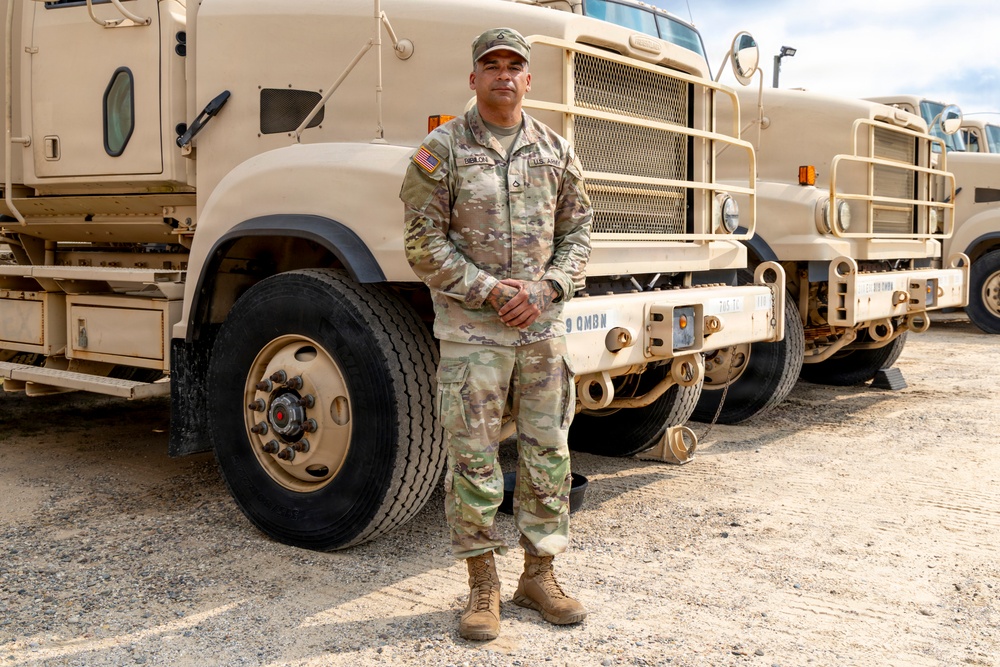 A U.S. Army soldier in OCP (Operational Camouflage Pattern) uniform stands at attention in front of large military tactical trucks. The vehicles are painted in desert tan and marked with '9 QMBN' (9th Quartermaster Battalion) identifiers. The setting appears to be a military motor pool or staging area on a gravel surface.