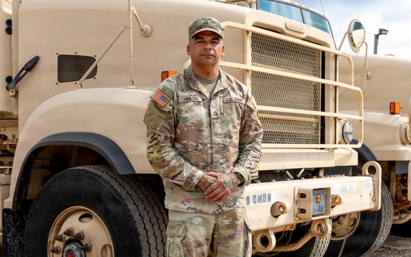 A U.S. Army soldier in OCP (Operational Camouflage Pattern) uniform stands at attention in front of large military tactical trucks. The vehicles are painted in desert tan and marked with '9 QMBN' (9th Quartermaster Battalion) identifiers. The setting appears to be a military motor pool or staging area on a gravel surface.