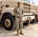 A U.S. Army soldier in OCP (Operational Camouflage Pattern) uniform stands at attention in front of large military tactical trucks. The vehicles are painted in desert tan and marked with '9 QMBN' (9th Quartermaster Battalion) identifiers. The setting appears to be a military motor pool or staging area on a gravel surface.