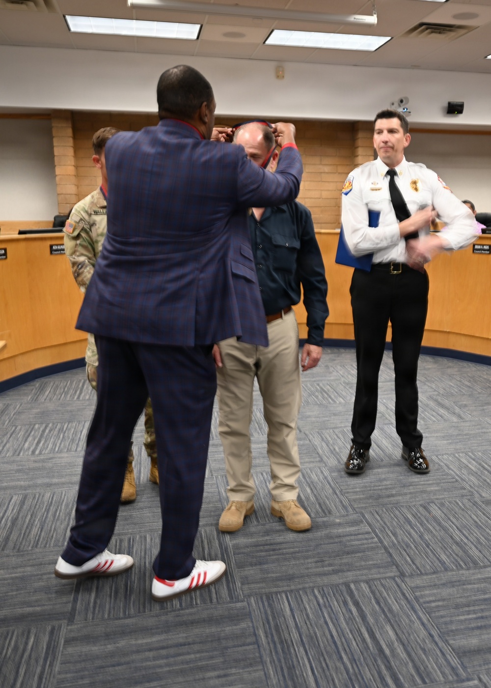 A formal ceremony appears to be taking place in what looks like a government council chamber or meeting room. A man in a blue plaid suit is placing or adjusting something on the head of another individual, while a uniformed fire department officer holding a blue folder stands nearby observing. A military personnel member in camouflage uniform is also visible in the background.