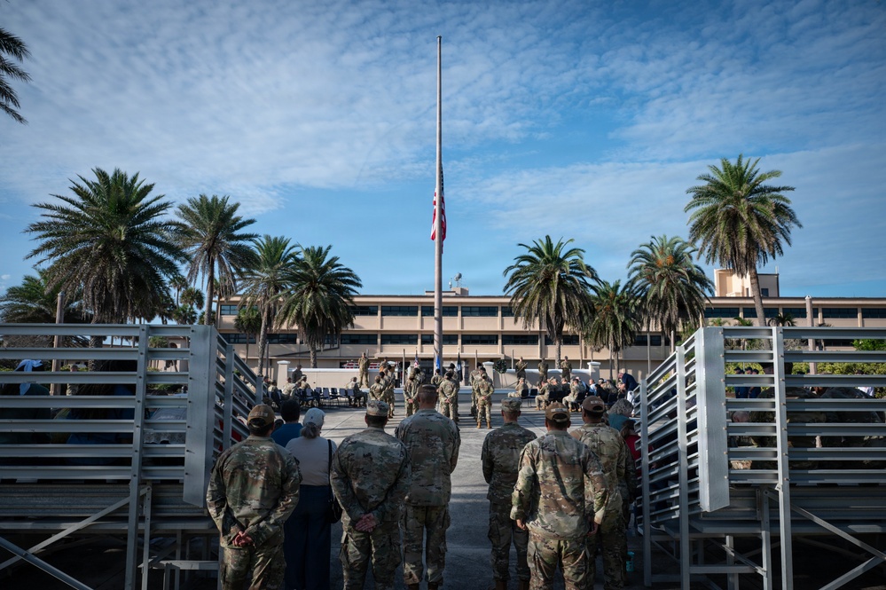 A group of military personnel in camouflage uniforms are gathered at what appears to be a military installation for a formal ceremony. The American flag is visibly flying at half-staff on a tall flagpole, suggesting a memorial or mourning observance. The setting includes a multi-story building flanked by palm trees, indicating a warm-climate military base.