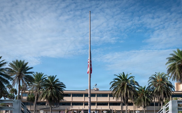 A group of military personnel in camouflage uniforms are gathered at what appears to be a military installation for a formal ceremony. The American flag is visibly flying at half-staff on a tall flagpole, suggesting a memorial or mourning observance. The setting includes a multi-story building flanked by palm trees, indicating a warm-climate military base.