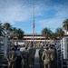 A group of military personnel in camouflage uniforms are gathered at what appears to be a military installation for a formal ceremony. The American flag is visibly flying at half-staff on a tall flagpole, suggesting a memorial or mourning observance. The setting includes a multi-story building flanked by palm trees, indicating a warm-climate military base.