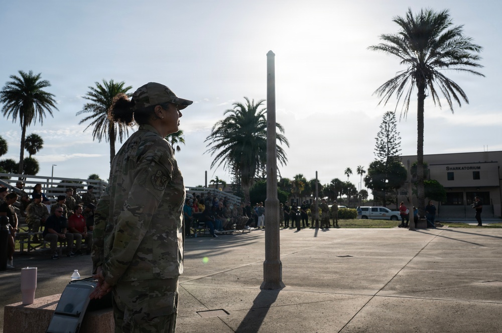 A female military service member in camouflage uniform stands at attention in an outdoor plaza, appearing to observe or participate in a formal military ceremony or gathering. A crowd of people, including other uniformed personnel and civilians, is visible in the background seated on bleachers and standing. The setting appears to be a coastal or recreational facility, evidenced by the 'Sharkatorium' building sign and palm trees in the background.