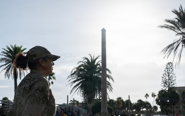 A female military service member in camouflage uniform stands at attention in an outdoor plaza, appearing to observe or participate in a formal military ceremony or gathering. A crowd of people, including other uniformed personnel and civilians, is visible in the background seated on bleachers and standing. The setting appears to be a coastal or recreational facility, evidenced by the 'Sharkatorium' building sign and palm trees in the background.