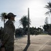 A female military service member in camouflage uniform stands at attention in an outdoor plaza, appearing to observe or participate in a formal military ceremony or gathering. A crowd of people, including other uniformed personnel and civilians, is visible in the background seated on bleachers and standing. The setting appears to be a coastal or recreational facility, evidenced by the 'Sharkatorium' building sign and palm trees in the background.