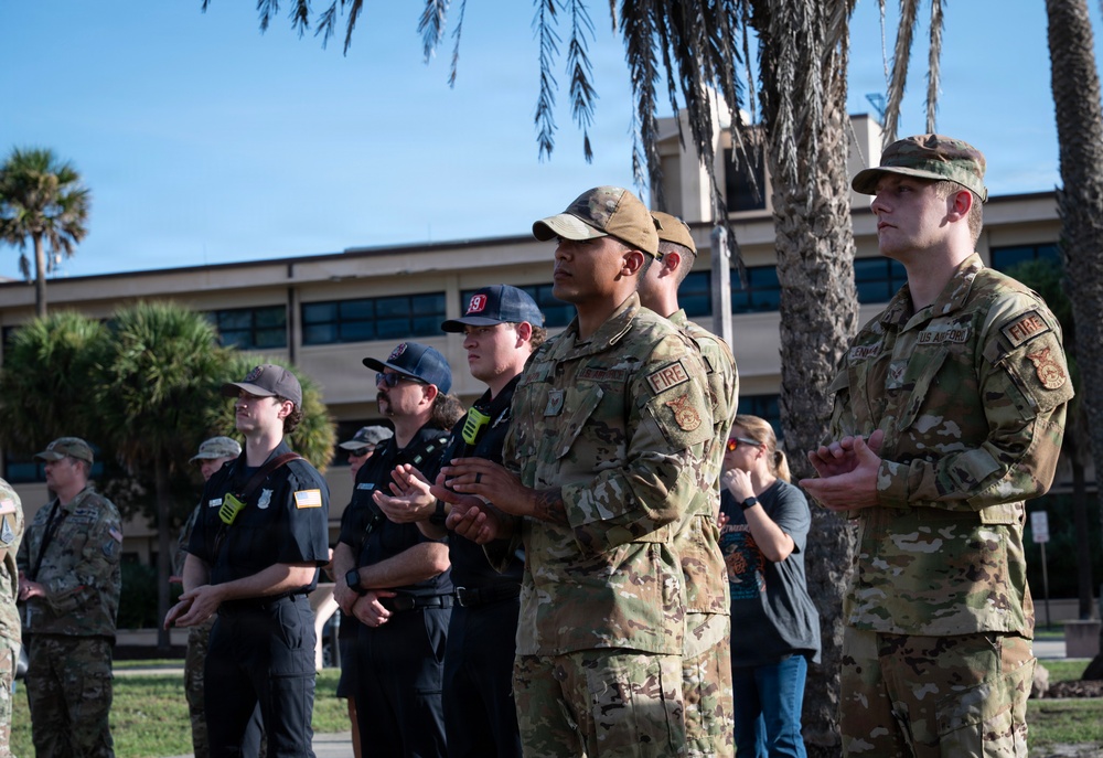 A group of U.S. Air Force personnel and civilian firefighters are gathered outdoors on what appears to be a military installation, likely during a ceremony or formal event. The individuals are standing at attention or applauding, wearing a mix of OCP (Operational Camouflage Pattern) uniforms and dark firefighter duty uniforms, several bearing 'FIRE' patches. The setting includes palm trees and a multi-story building consistent with a military base in a warm-climate region.