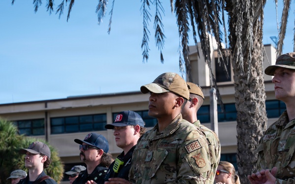 A group of U.S. Air Force personnel and civilian firefighters are gathered outdoors on what appears to be a military installation, likely during a ceremony or formal event. The individuals are standing at attention or applauding, wearing a mix of OCP (Operational Camouflage Pattern) uniforms and dark firefighter duty uniforms, several bearing 'FIRE' patches. The setting includes palm trees and a multi-story building consistent with a military base in a warm-climate region.